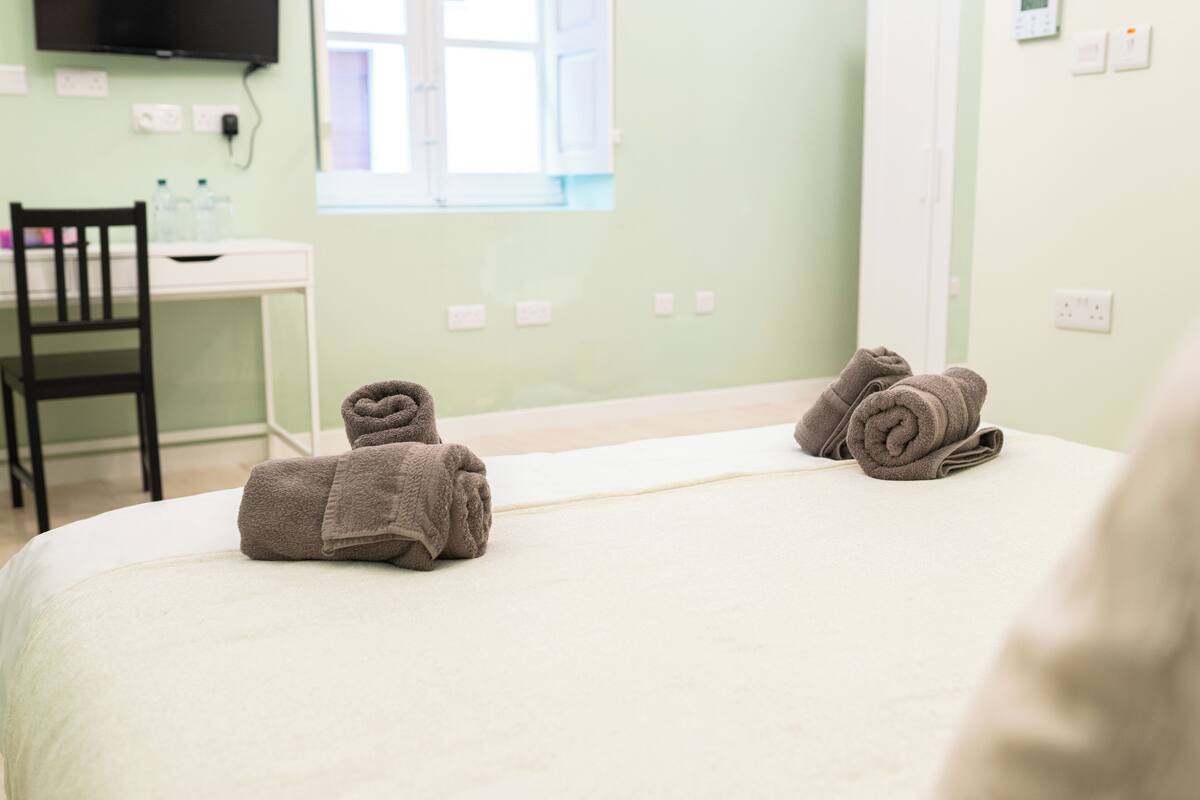A hotel room with a bed, two rolled towels, a desk, and a chair.
