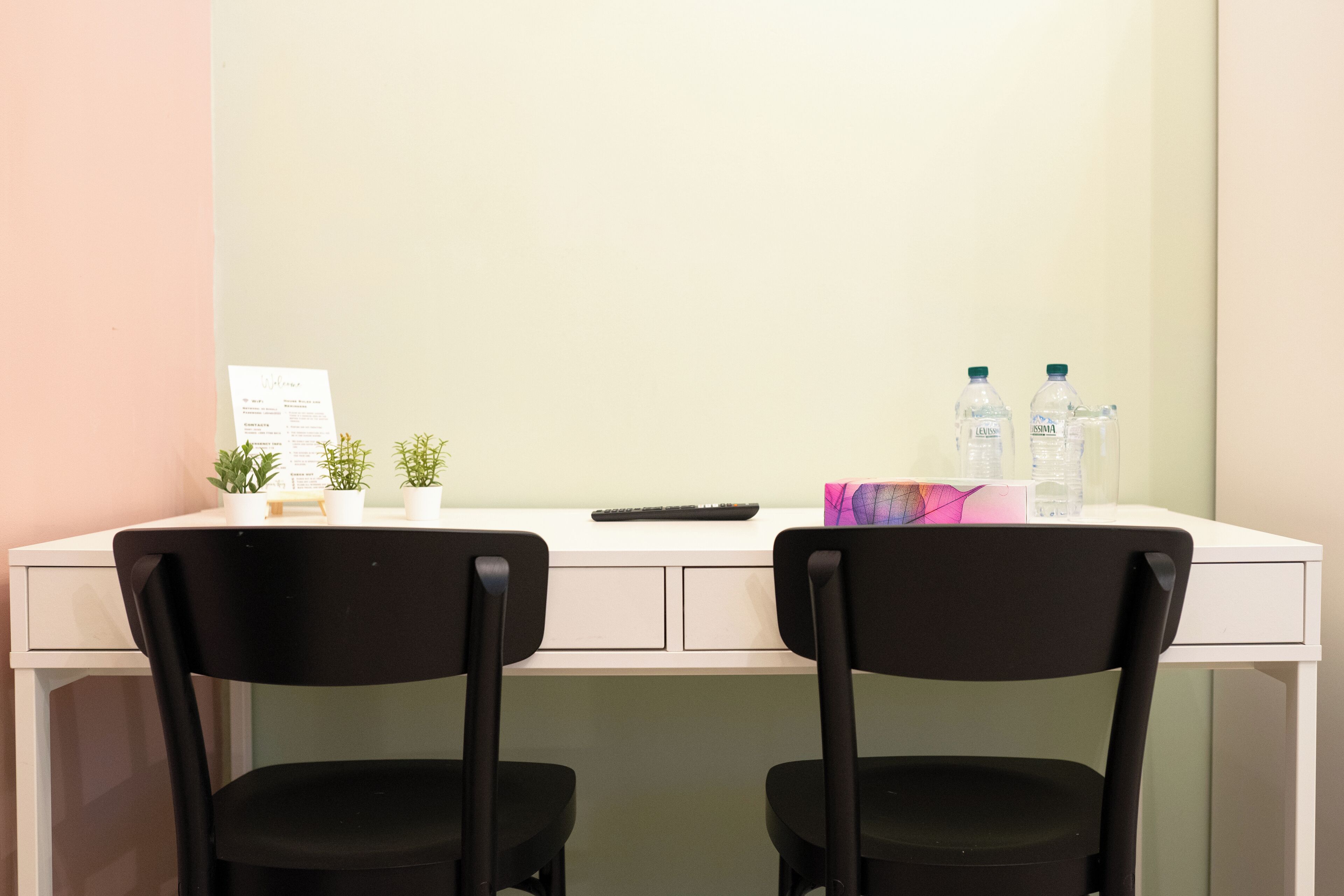 A white desk with two black chairs, a keyboard, a small potted plant, a water bottle, and a box of tissues.