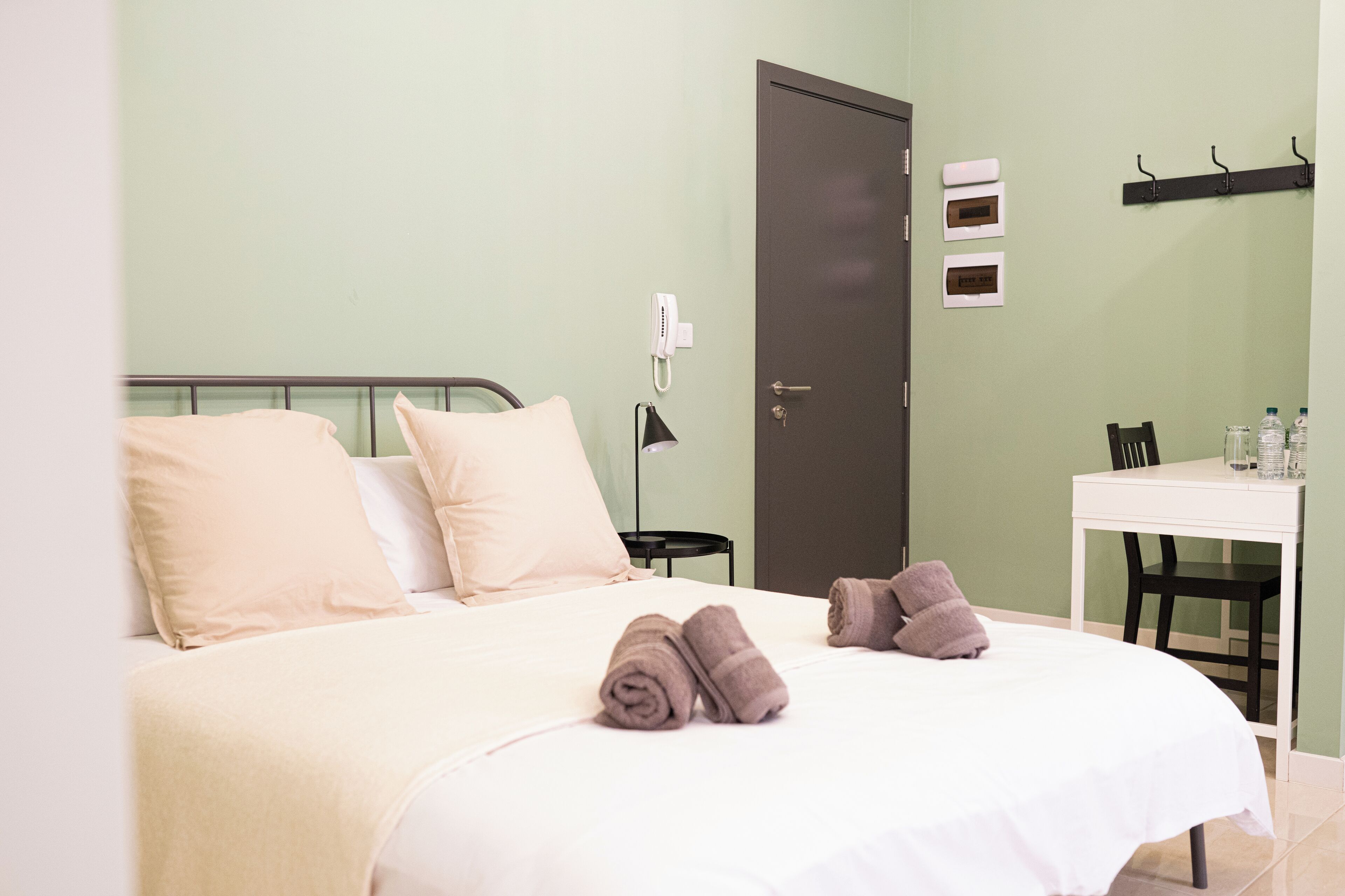 A neatly made bed with pillows and rolled towels, a bedside table with a lamp, a door, and a small table with bottles and glasses.
