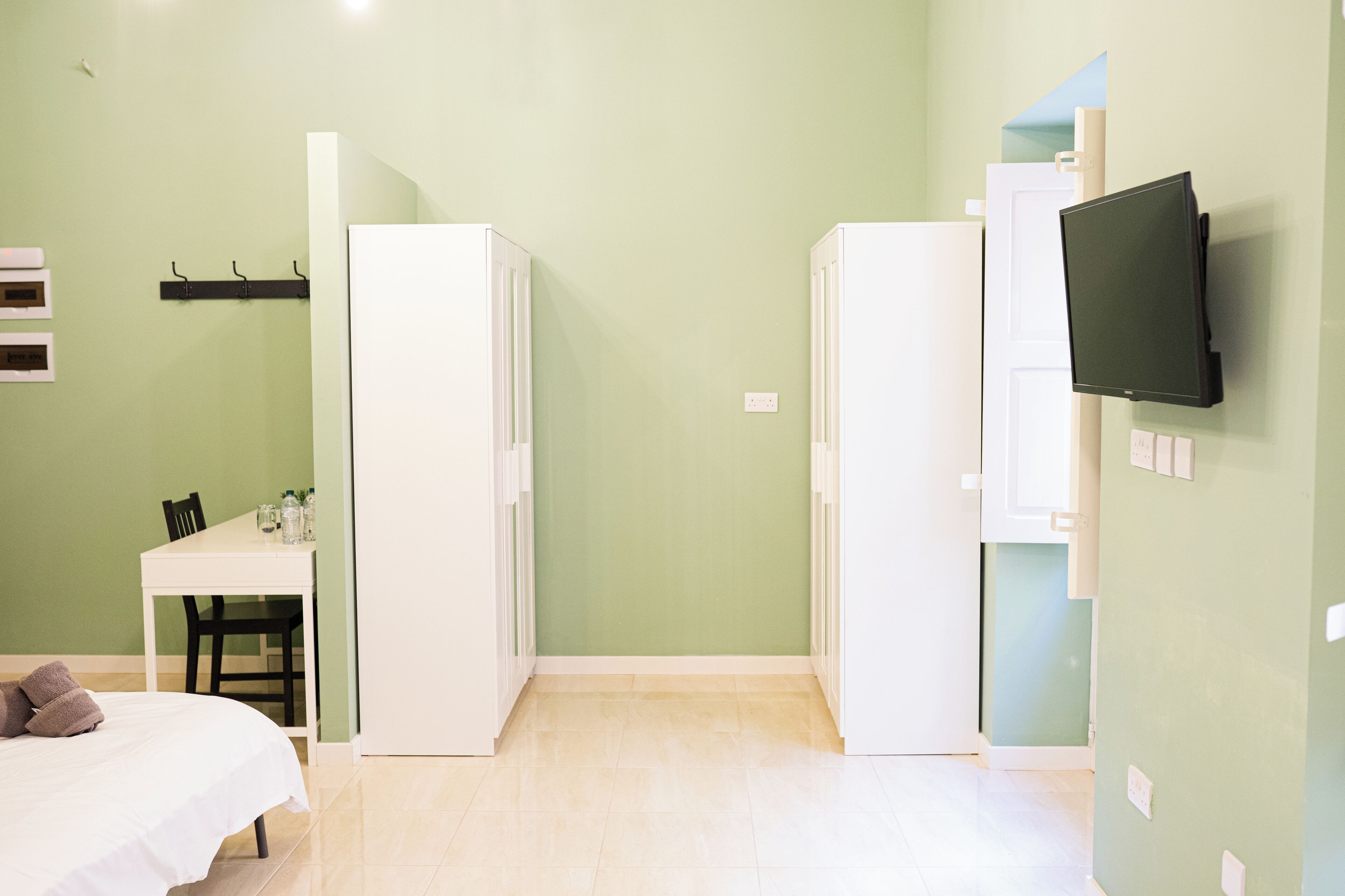 A room with a bed, a desk, two white wardrobes, a flat-screen TV mounted on the wall, and a small table with bottles.