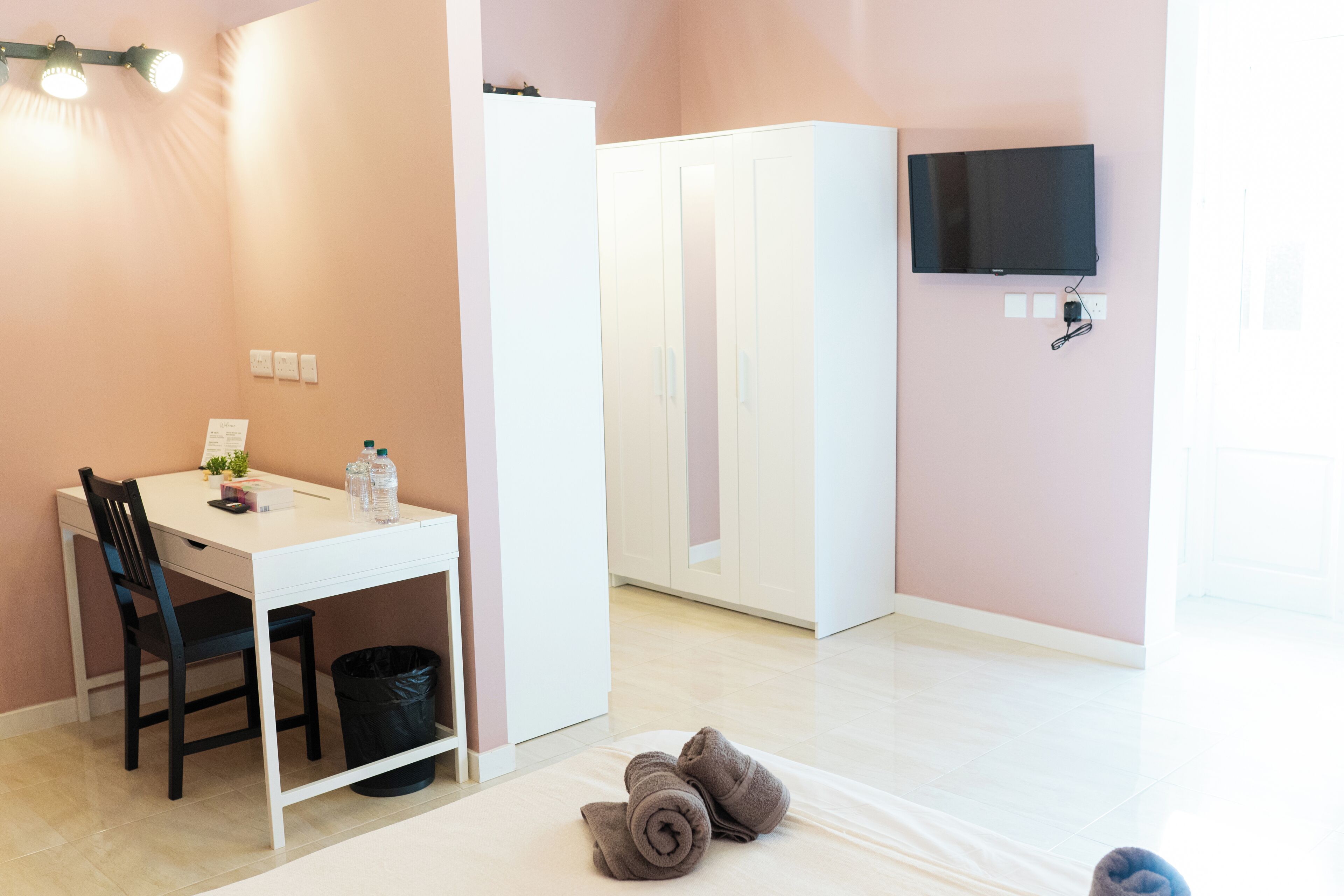 Een kamer met een wit bureau, een stoel, een kledingkast, een aan de muur bevestigde televisie en een prullenbak.