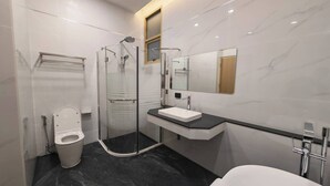 Un baño moderno con ducha de vidrio, inodoro blanco, suelo de mármol negro, lavamanos blanco y un espejo grande.