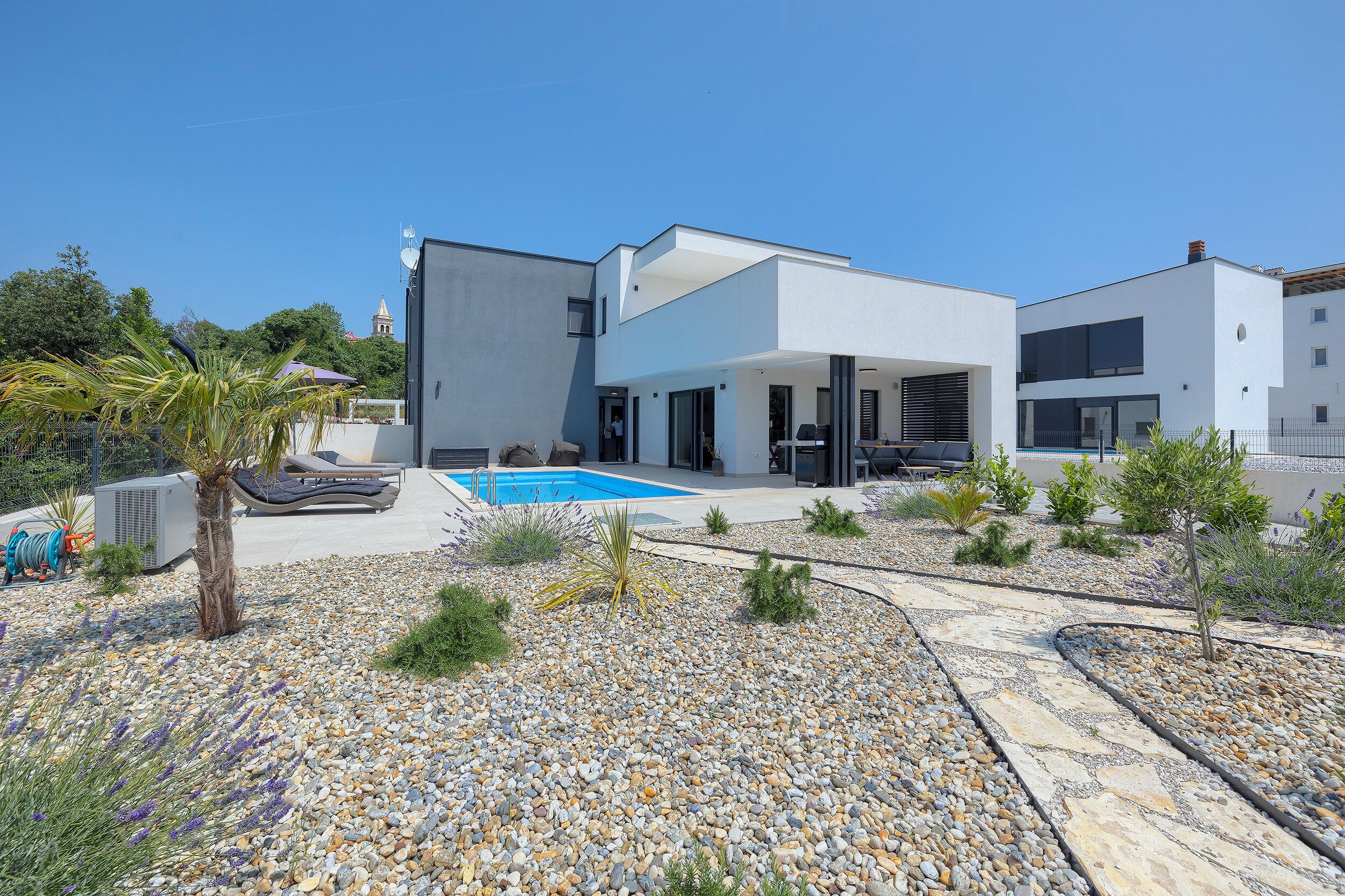 A modern two-story house with a swimming pool, surrounded by a landscaped garden with various plants and a pebble path.