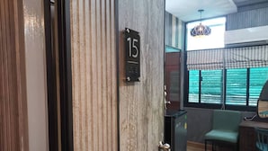 Une pièce avec une porte en bois portant le numéro 15, un petit meuble noir, une chaise verte et une fenêtre donnant sur la ville.