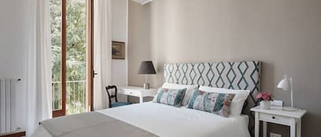 A bedroom with a large bed, a nightstand, a lamp, and a view of greenery through a window.