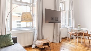 Huoneessa on sohva, pieni pöytä tuoleineen, lattiavalaisin ja seinälle asennettu televisio.