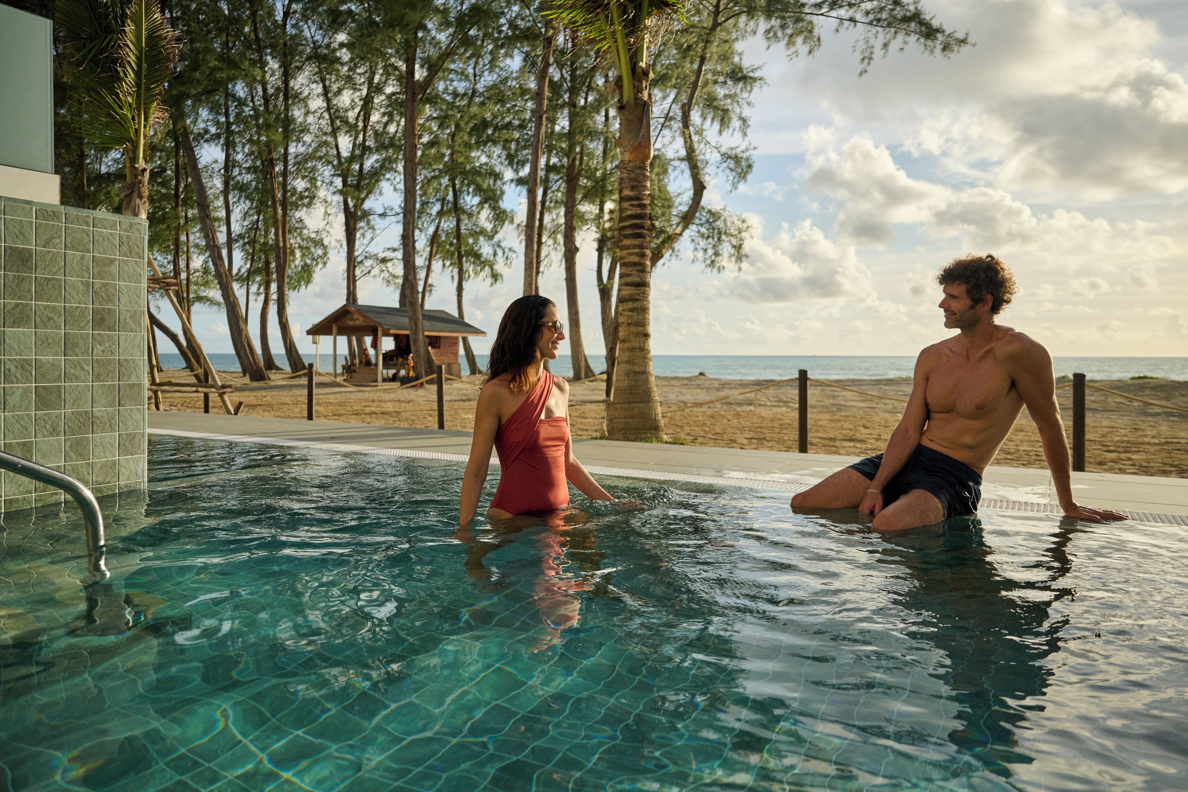 A swimming pool with clear water, surrounded by palm trees and a beach view.