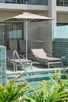 Zona de piscina con sillones de descanso y una mesa, una sombrilla y una sala acristalada al fondo.