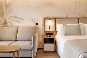 A hotel room with a sofa, a bed, a nightstand with a lamp, and a wooden table. - Hotel Riu Palace Phuket - All Inclusive (Mai Khao)