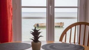 Uitzicht vanuit een raam, met een houten stoel en een tafel, op een rustige zee en een betonnen constructie.