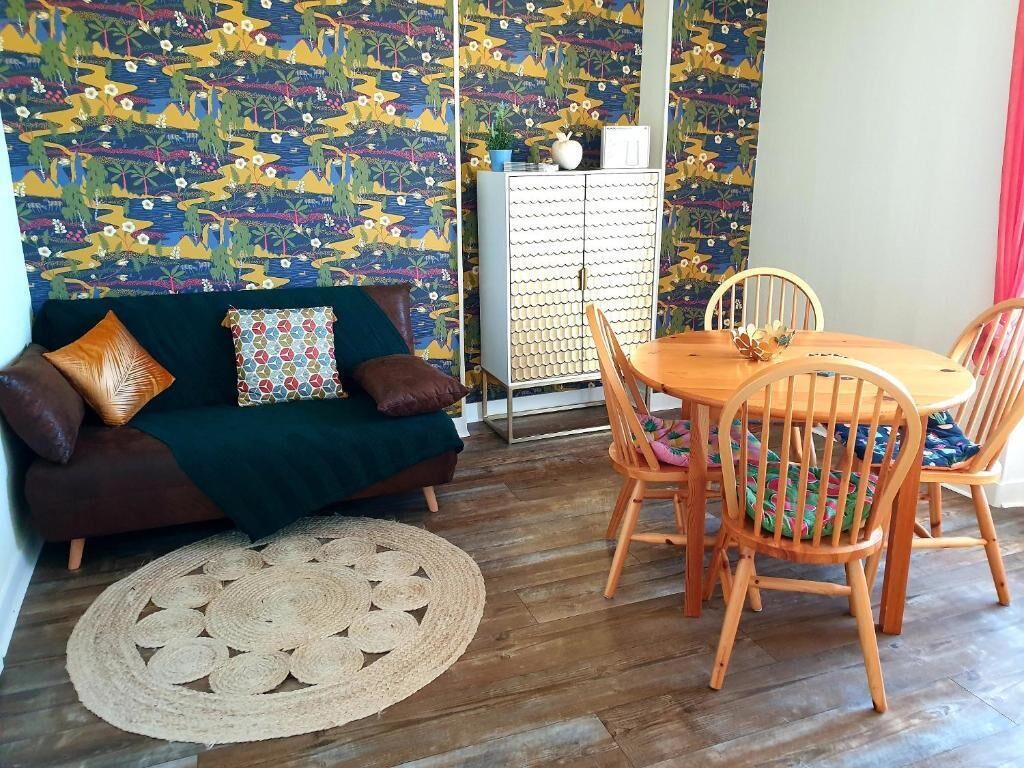 Un salon chaleureux avec un papier peint à motifs, un canapé vert foncé agrémenté de coussins à motifs, une table à manger ronde en bois avec des chaises, et un tapis tissé.