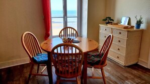 Een eethoek met een ronde houten tafel en vier stoelen, een klein kastje met decoratieve spullen, en uitzicht op het strand door een raam.