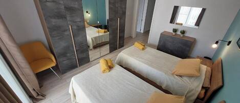 A modern hotel room with two beds, a desk, a mirror, and a bathroom visible through a sliding door.