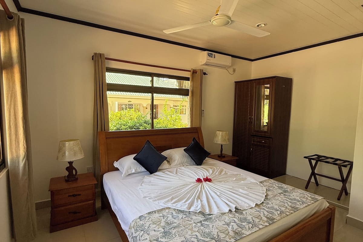 A neatly made bed with a white and patterned bedspread, a wooden headboard, and two black pillows. There is a nightstand with a lamp, a ceiling fan, and a window with curtains.