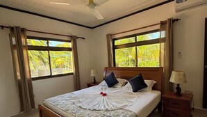 A well-lit bedroom with a wooden bed, two bedside tables, a ceiling fan, and large windows with curtains.