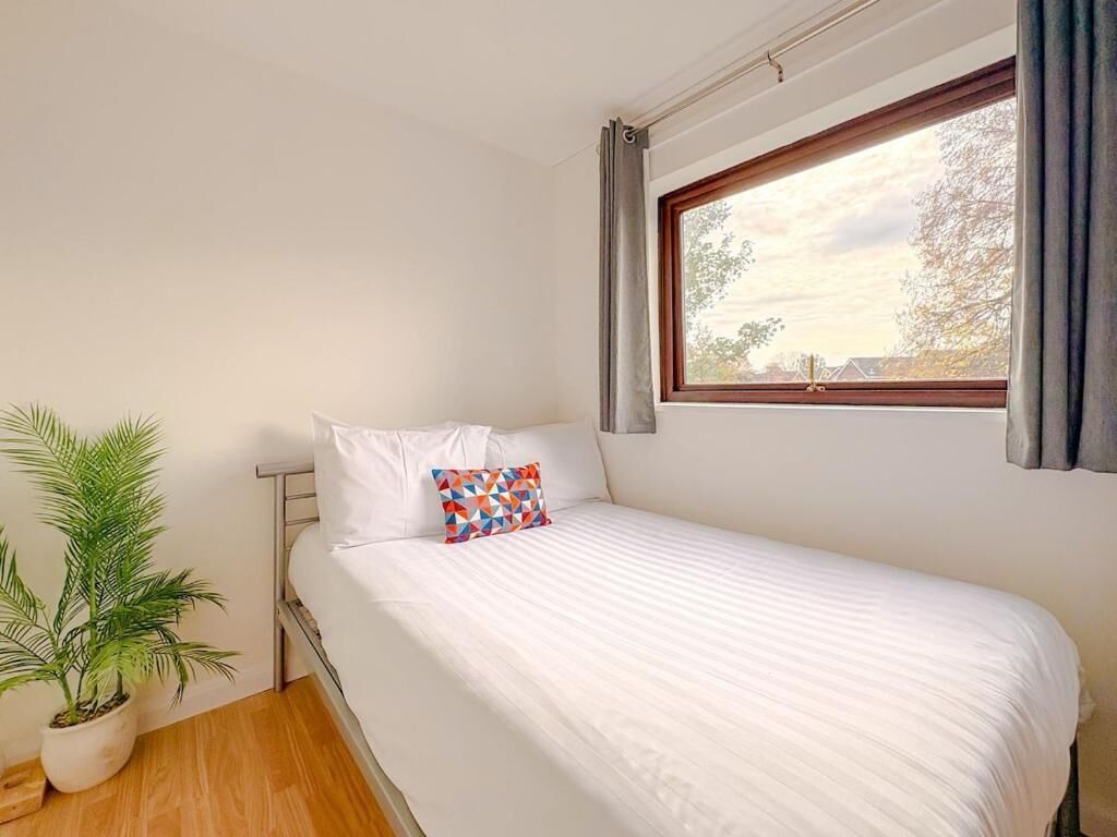 Una cama individual con ropa blanca y un cojín colorido, una planta en maceta y una ventana con cortinas.