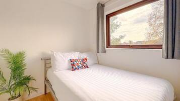Una cama individual con ropa blanca y un cojín colorido, una planta en maceta y una ventana con cortinas.