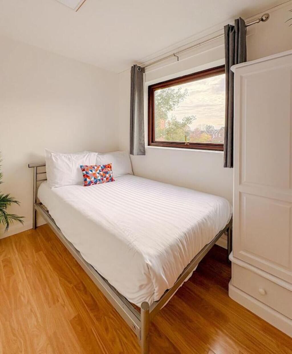 A single bed with white bedding, a colorful pillow, a window with curtains, and a wooden floor.