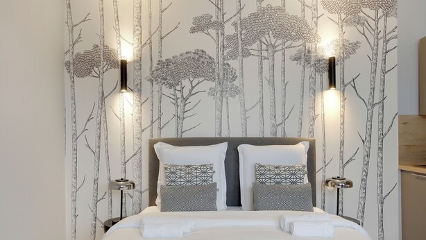 A neatly made bed with white linens and geometric patterned pillows, set against a wall with a nature-themed wallpaper and two wall-mounted light fixtures. - Appartements rue Garnier (Neuilly-sur-Seine)