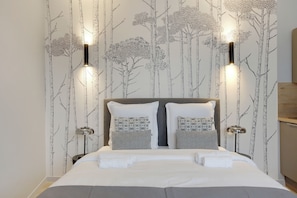 Een net opgemaakt bed met witte lakens en kussens met een geometrisch patroon, geplaatst tegen een muur met behang met een natuurthema en twee wandlampen.