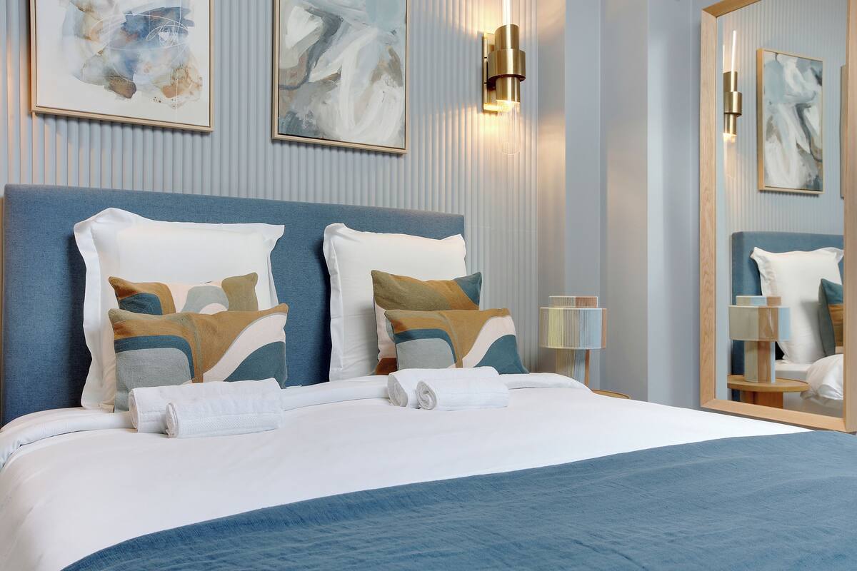 A neatly made bed with a blue headboard, white and patterned pillows, and a rolled white towel. There are two wall-mounted light fixtures and framed abstract art above the bed.