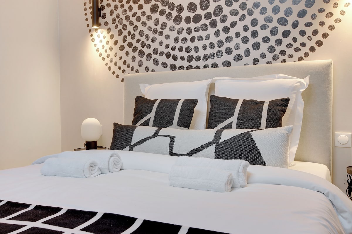 A neatly made bed with white linens and black and white patterned pillows, a wall with a circular dot pattern, and a bedside lamp.