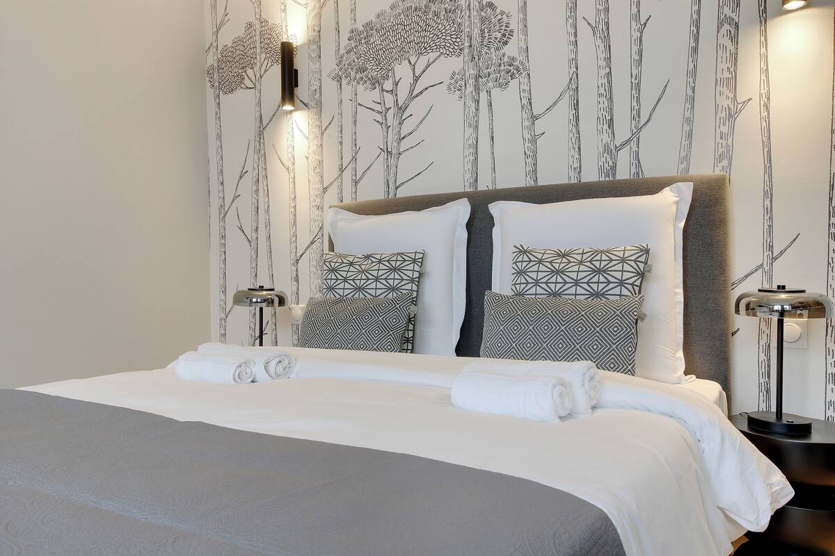 A neatly made bed with white linens and geometric patterned pillows, a bedside table with a lamp, and a wall with a nature-themed wallpaper.