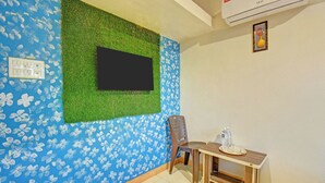 Ein Zimmer mit an der Wand befestigtem Fernseher, einem Sessel, einem kleinen Tisch mit Wasserflasche und einem Thermostat.