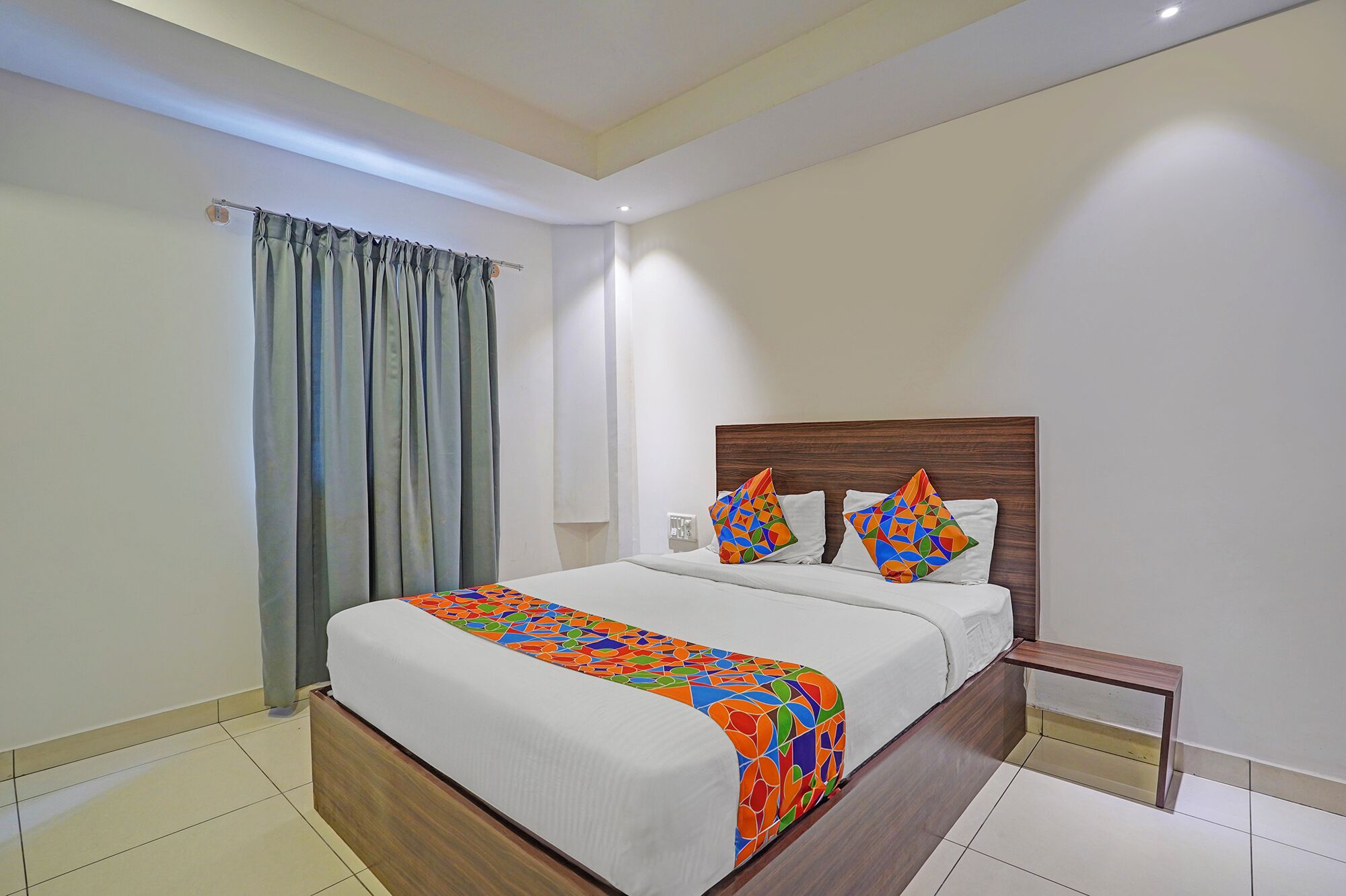 A hotel room with a wooden bed frame, white bedding, and colorful geometric pillows.
