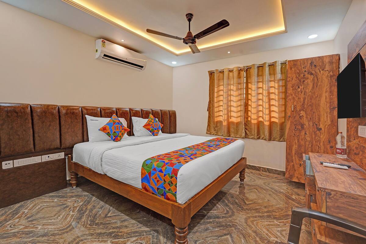 A hotel room with a wooden bed, a ceiling fan, an air conditioner, a window with curtains, and a desk with a chair.