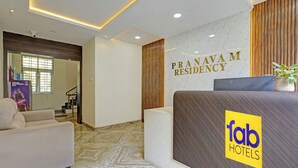 Zona de recepción de un hotel con suelo de mármol, mostrador de recepción con un logotipo, una escalera y una ventana con persianas.