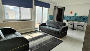 A modern living room with a sofa, a small table, and a chair. There are large windows with blinds, a door, and a wall with a decorative tile border. - Fabhotel Ekab Hospitality (Pune)