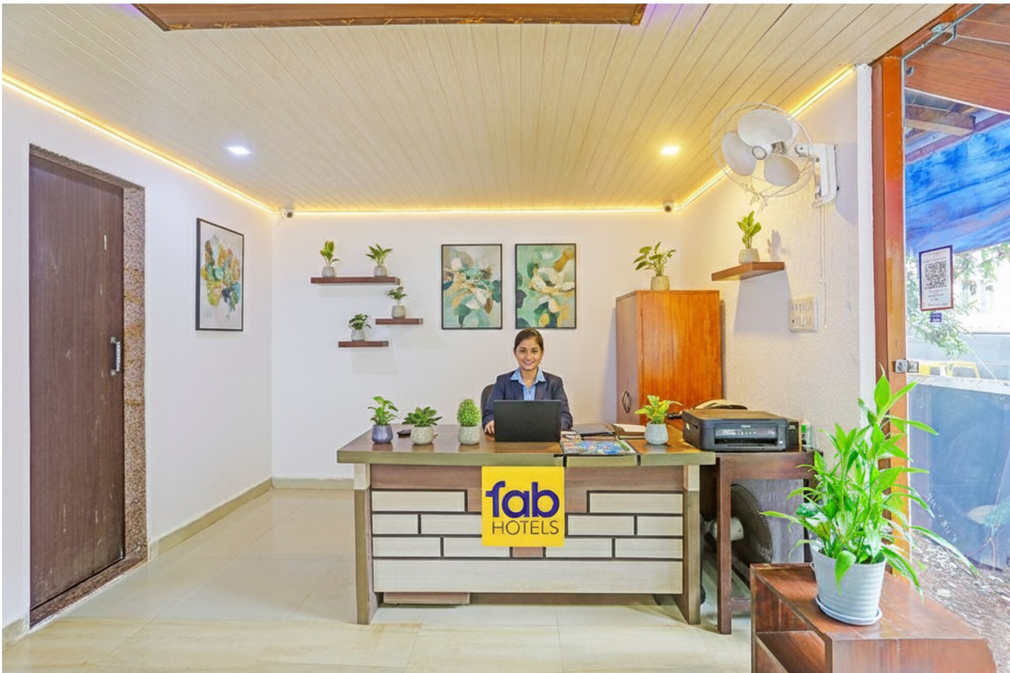 A hotel reception desk with a receptionist, a computer, and a fax machine. There are several potted plants on shelves and a wooden cabinet.