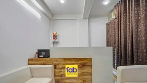 A hotel reception area with a wooden reception desk, a sign for 'fab hotels', two sofas, a small shelf with a bottle, and a curtain. - Fabhotel Aakarshan Residency (New Delhi)