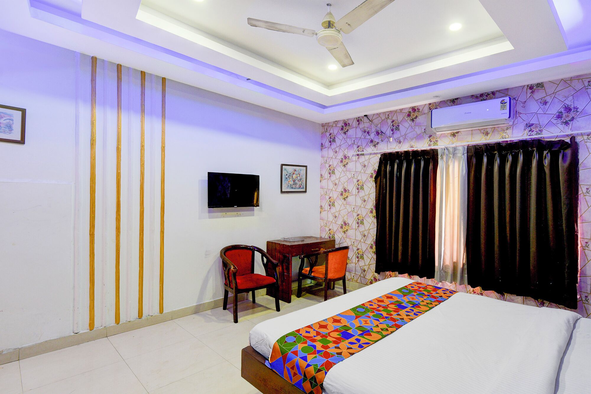 A hotel room with a bed, two chairs, a desk, a television, and an air conditioning unit.