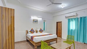A hotel room with a bed, two chairs, a table, a ceiling fan, an air conditioning unit, and a wooden door. - Fabhotel Casa Residency (Noida)