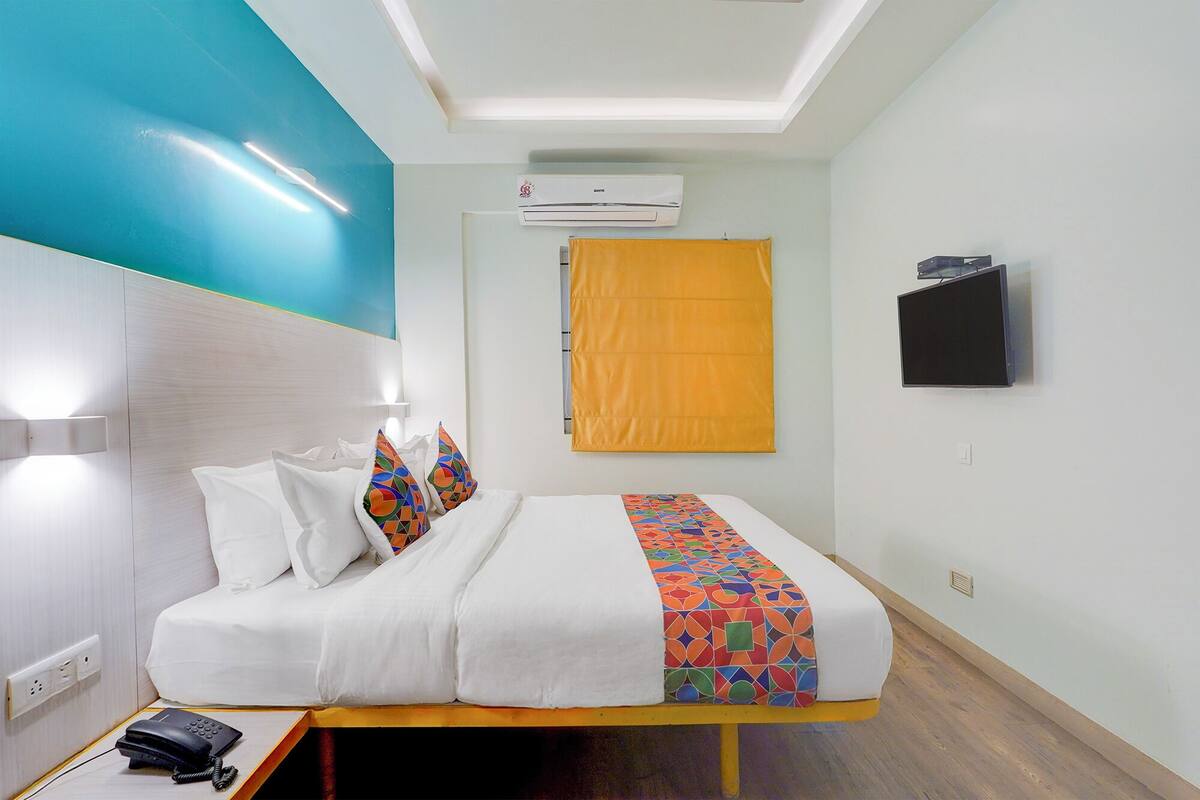 A hotel room with a bed, a bedside phone, an air conditioning unit, a television, and a window with a yellow curtain.