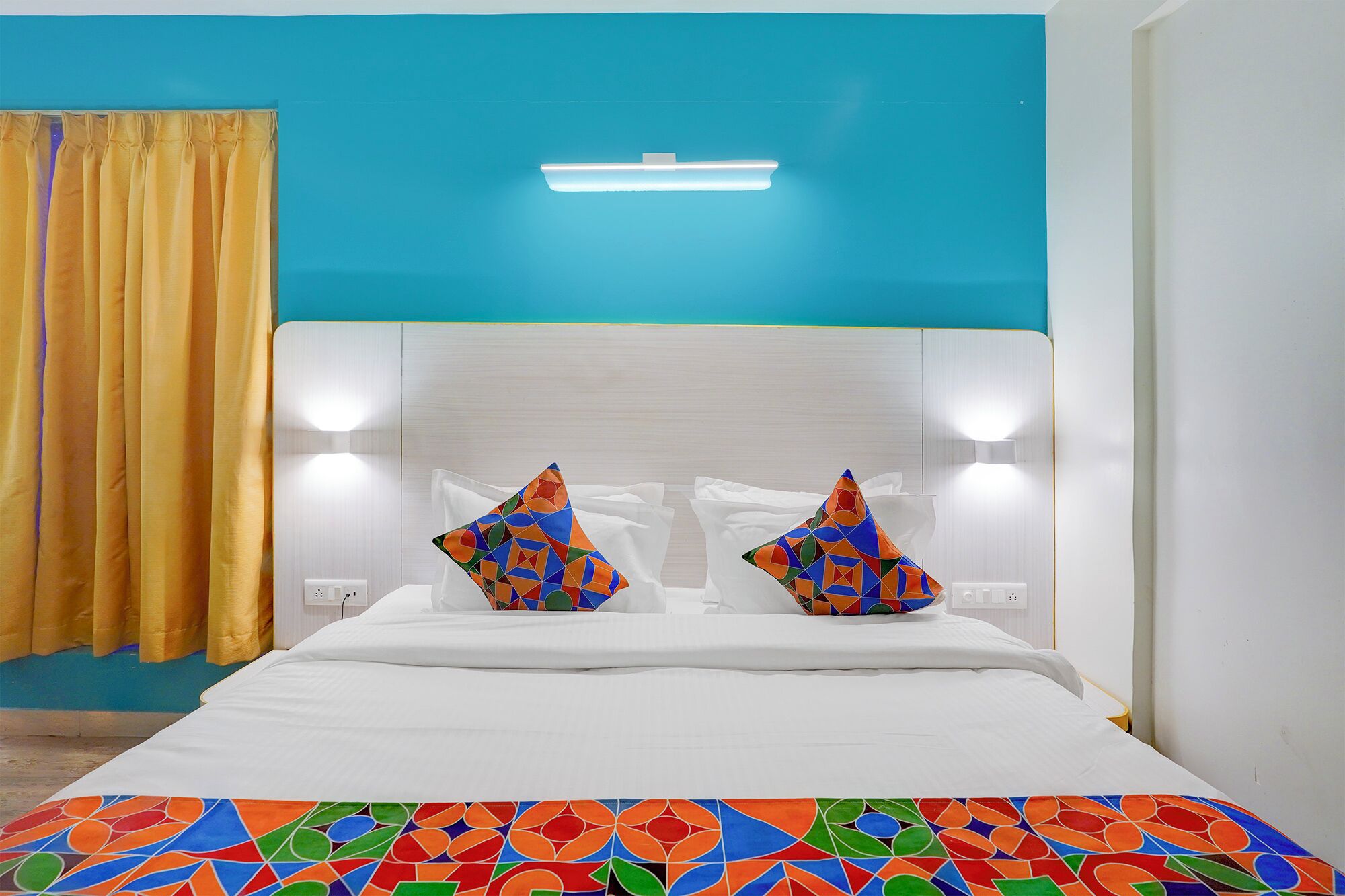 A hotel room with a bed, two decorative pillows, a ceiling light, and a yellow curtain.