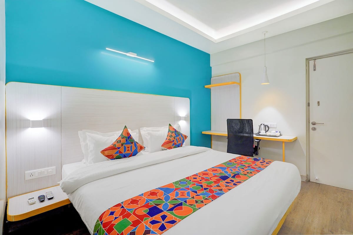 A hotel room with a bed, a bedside table with a lamp, a small desk with a kettle, and a door.