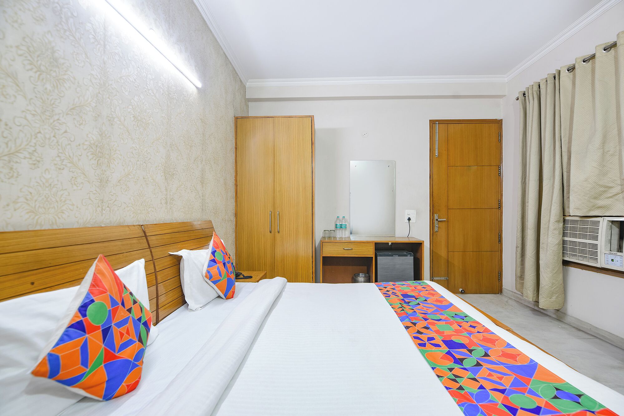 A hotel room with a bed, a wooden wardrobe, a desk with a mirror, and a window with curtains.