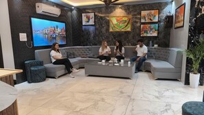 A modern lounge area with a grey sectional sofa, a coffee table, and a flat-screen TV displaying a cityscape. - Fabhotel Yatri Nivas (Varanasi)