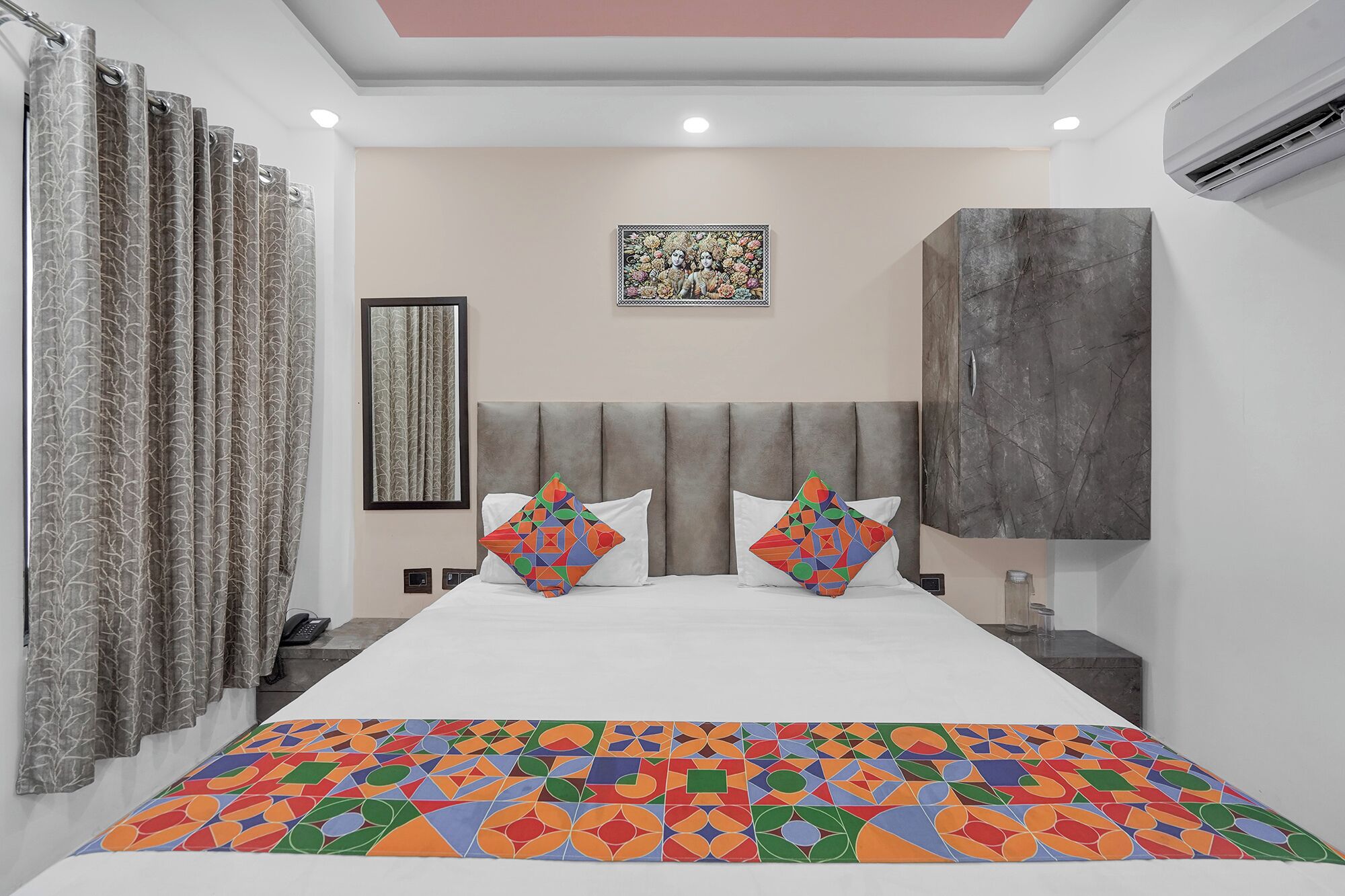 A modern hotel room with a large bed, a headboard, a framed artwork, an air conditioning unit, and a patterned bedspread.
