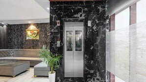 A modern lobby with a marble wall, a grey sofa, a potted plant, and a digital elevator display. - Fabhotel Yatri Nivas (Varanasi)
