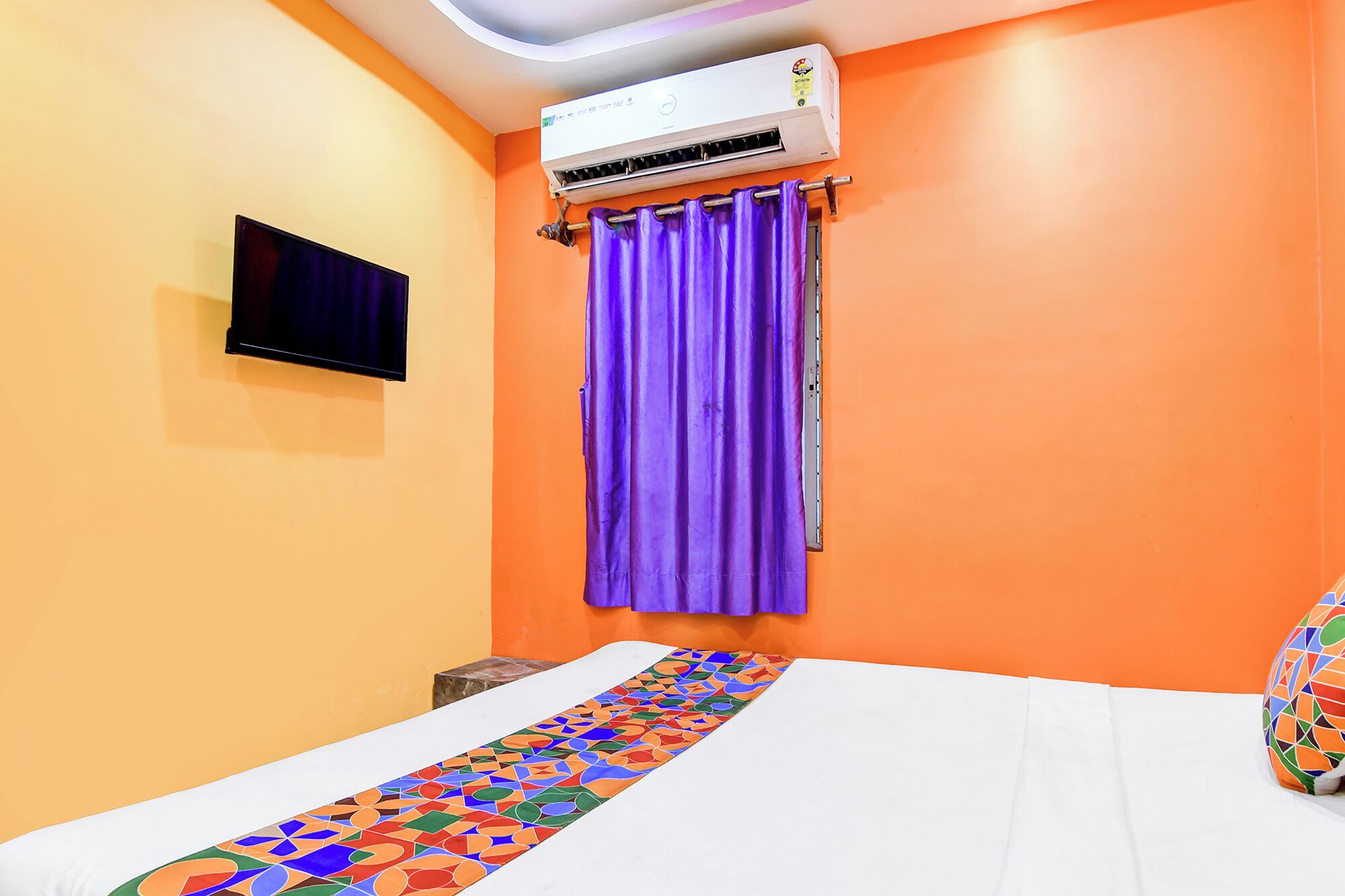Una stanza con condizionatore, un letto con copriletto fantasia, una televisione a parete e una finestra con tende viola.