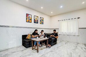 Una sala de estar moderna con piso de mármol, un sofá negro, una mesa de centro de madera y tres personas sentadas usando dispositivos electrónicos.