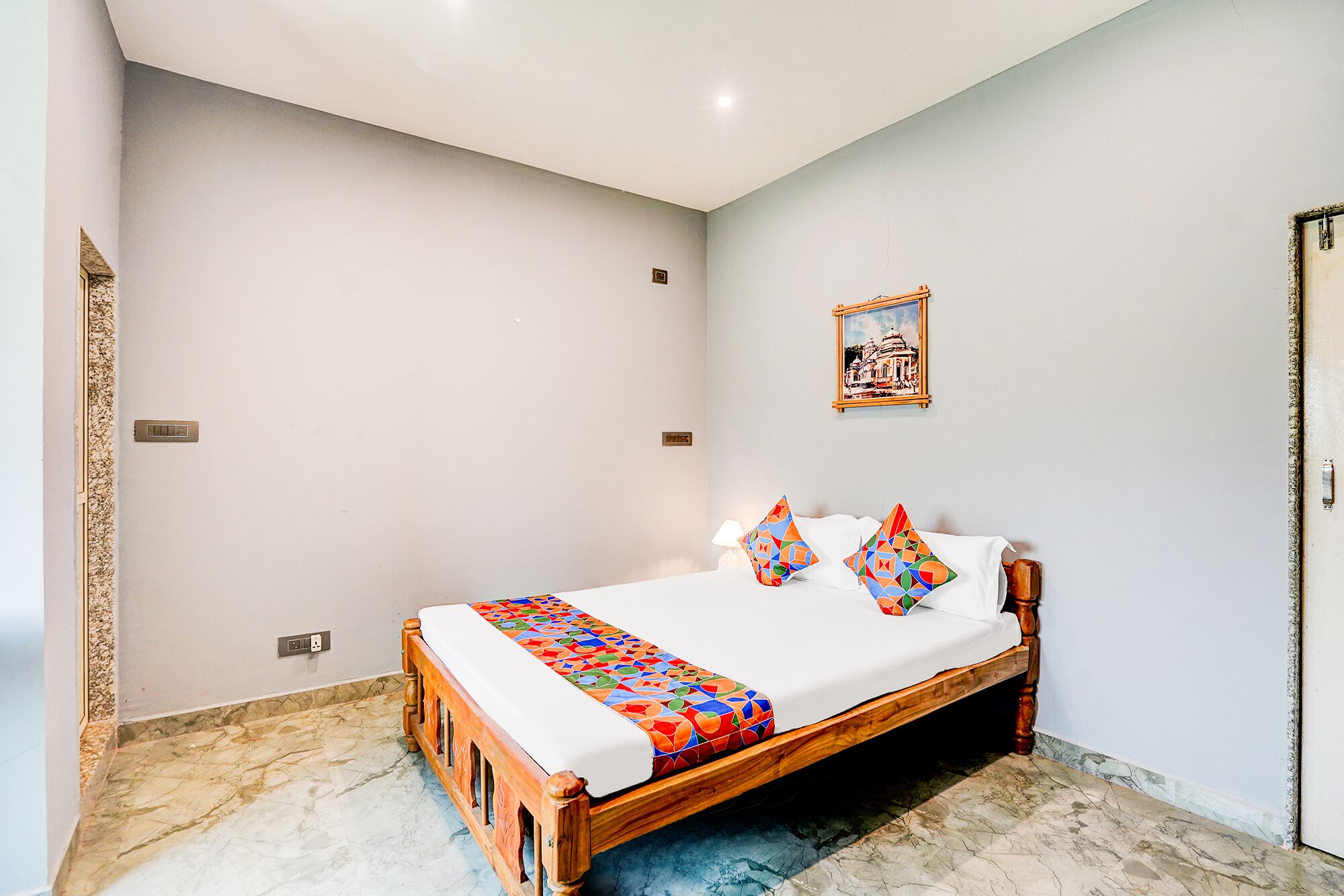 Una cama bien hecha con una colcha de diseño geométrico y colorido, dos almohadones decorativos, un cuadro enmarcado en la pared y un marco de madera para la puerta.