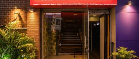 A entrada do hotel Sunset Avenue, com porta de vidro, escadaria que leva para dentro e plantas em vasos lá fora.