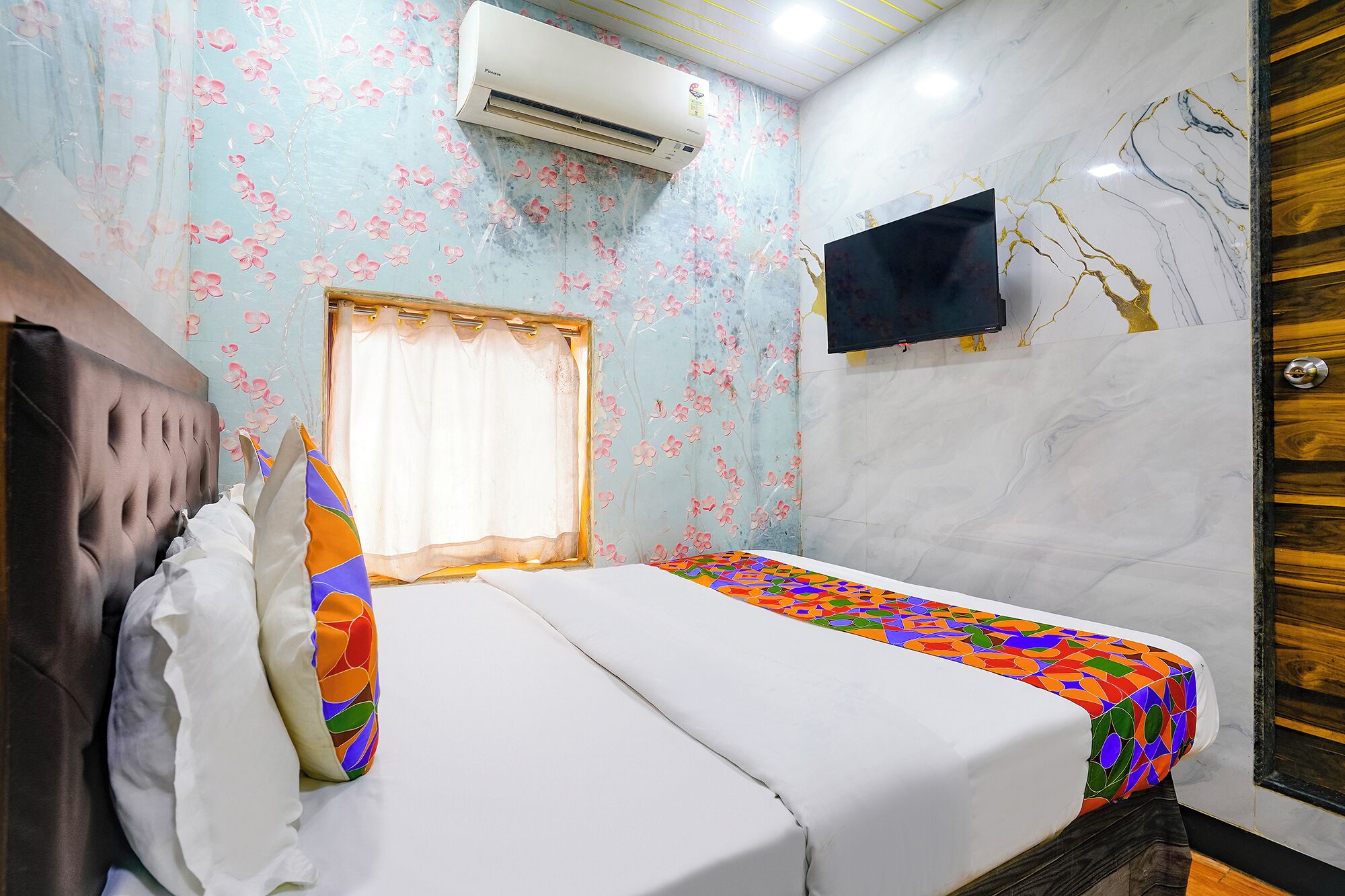 A hotel room with a bed, a window with a curtain, an air conditioning unit, and a television mounted on the wall.