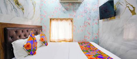 A hotel room with a bed, a window with curtains, an air conditioning unit, and a flat-screen TV mounted on the wall.