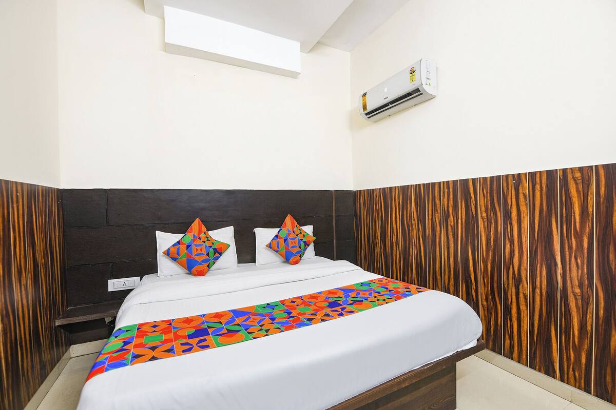 A hotel room with a bed, a wooden headboard, a white wall, and an air conditioning unit.
